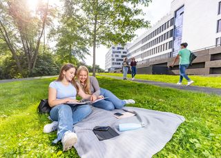 Zwei Studentinnen sitzen auf der Wiese am Gesundheitscampus und schauen in einen Ordner mit Unterlagen hinein.