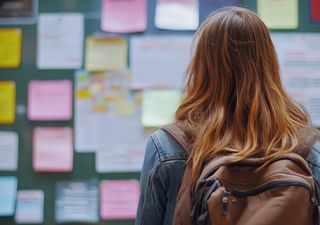 Dekoratives Bild. Im vordergrund steht eine Person mit langen Haaren und Rucksack, die von hinten zu sehen ist. Der Hintegrund ist unscharf, dort ist Wand mit vielen buten Zetteln und Post-Its zu sehen.