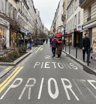 Fußverkehr in Paris Schmale Geschäftsstraße in Paris, auf der Fahrbahn sind große Beschriftungen angebracht, aus denen hervorgeht, dass Zufussgehende bevorrechtigt sind. Die Straße wird in ganzer Breite durch Passanten genutzt, gelegentlich parkt ein Auto vor einem Laden.